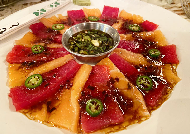 A platter of tuna and salmon sashimi arranged in a circular pattern, topped with fresh greens and a small bowl of ponzu dipping sauce in the center.
