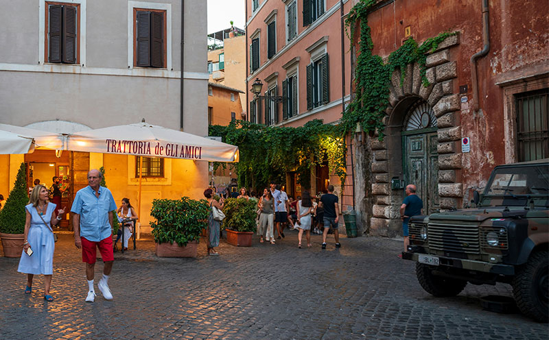 A view of a narrow, cobbled street in the Trastevere neighborhood of Rome, with an Italian military jeep parked near pedestrians and ivy-covered buildings.