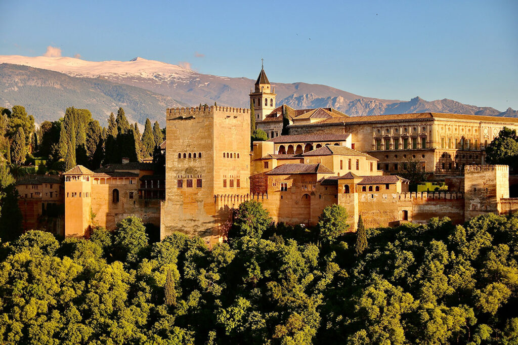 The Alhambra palace in Granada, Spain, standing on a wooded hill against a backdrop of mountains and a blue sky.