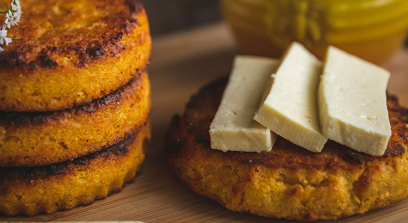 Two golden-brown Arepas de Choclo (Colombian sweet corn cakes) served warm with thick slices of fresh white cheese on a wooden board.