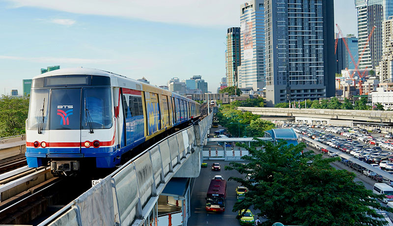 A BTS Skytrain moving along an elevated track above the Bangkok city streets during the day.