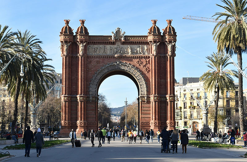 The Arc de Triomf in Barcelona, a red brick triumphal arch, viewed from the promenade with palm trees and people.
