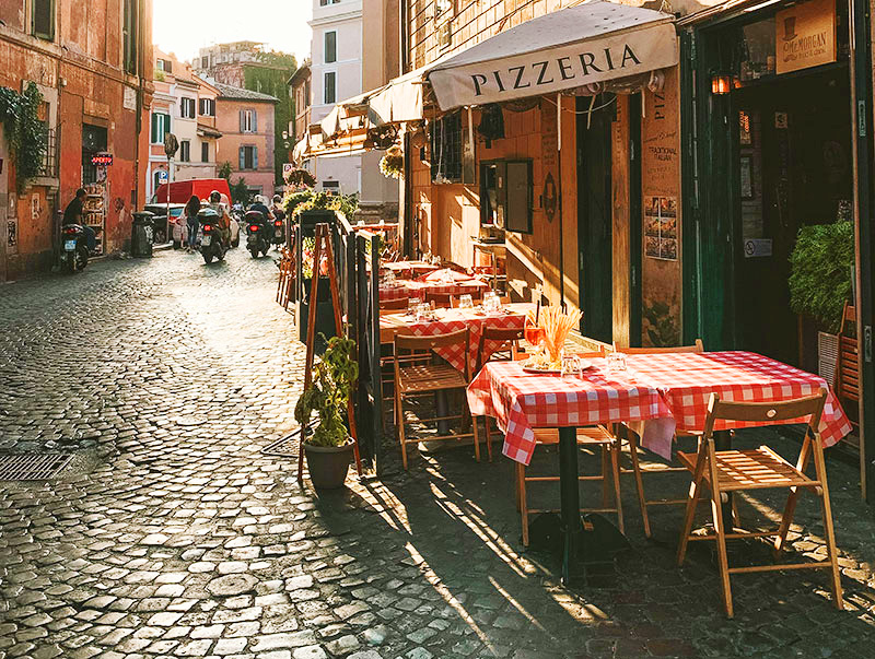 A narrow cobblestone street in Rome's Trastevere neighborhood lined with outdoor patio tables at a pizzeria, decorated with ivy and warm lighting.