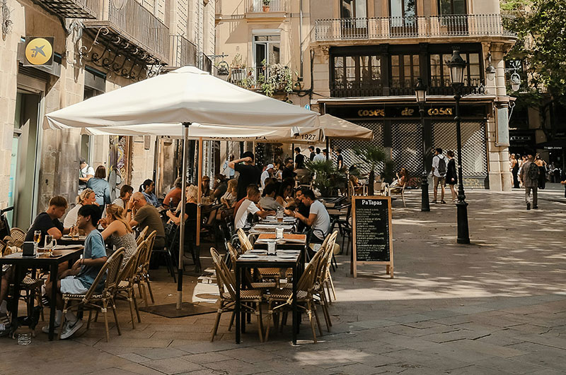Outdoor cafe in Barcelona with people seated at tables under white umbrellas on a sunny day.