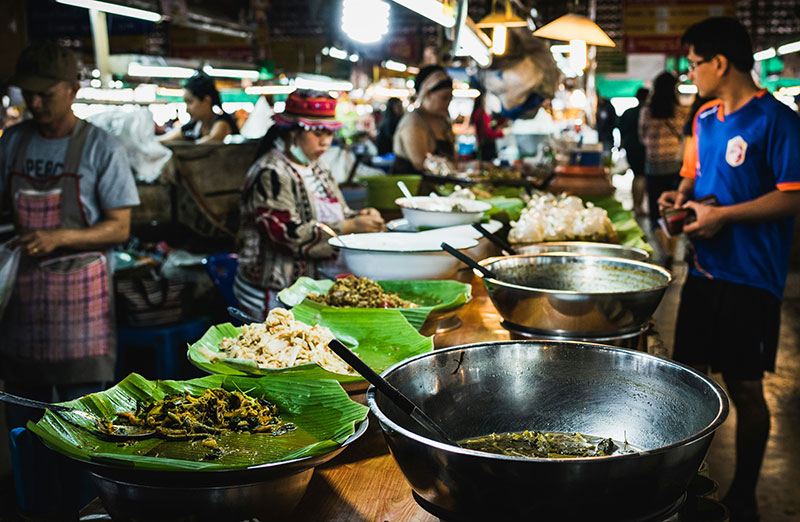 Vendors behind a counter at a busy Thai market serving various prepared dishes displayed in large silver bowls lined with banana leaves.