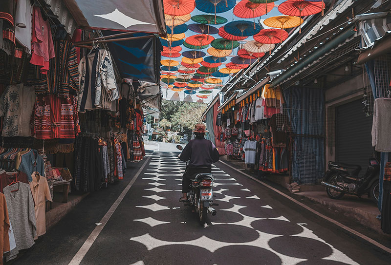 A person riding a motorbike down a narrow street covered by hundreds of hanging red, yellow, and blue umbrellas, creating geometric shadows on the road.