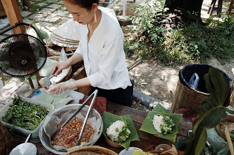 A street food vendor carefully plating portions of steamed rice and fresh herbs onto traditional banana leaves at an outdoor stall. Alt Tag: A woman wearing a white shirt and plastic gloves preparing food at a wooden table, featuring fresh cilantro, fried garlic, and rice served on banana leaves.