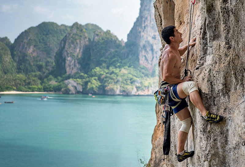 A man rock climbing on a steep limestone cliff face overlooking the turquoise ocean and a longtail boat at Tonsai Beach, Krabi, Thailand.