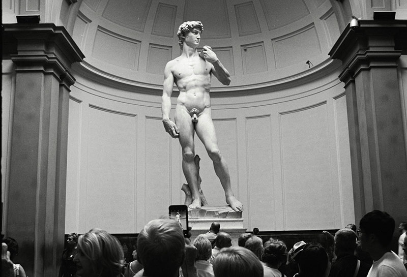 A low-angle view of Michelangelo's David statue in the Accademia Gallery, towering over a crowd of tourists.