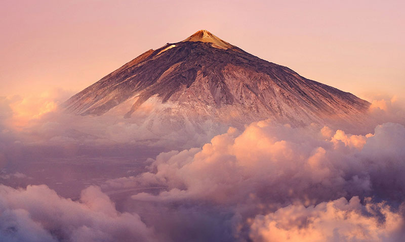 The snow-capped peak of El Teide volcano in the Canary Islands, rising above a layer of clouds with a rocky foreground.