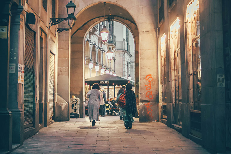 Street view in the Gothic Quarter of Barcelona, looking through an archway, with two people walking away under old stone buildings.
