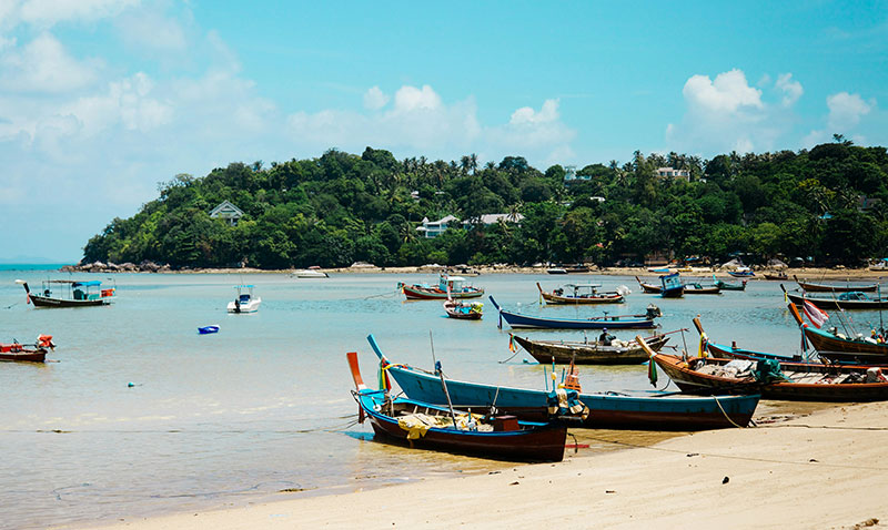 Traditional Thai longtail boats parked on a white sand beach in Koh Samui with green hills in the background.