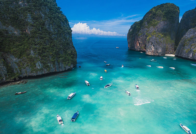 Aerial view of the turquoise waters of Maya Bay, Phi Phi Islands, dotted with numerous white speedboats and framed by towering limestone cliffs covered in lush green vegetation under a blue sky.