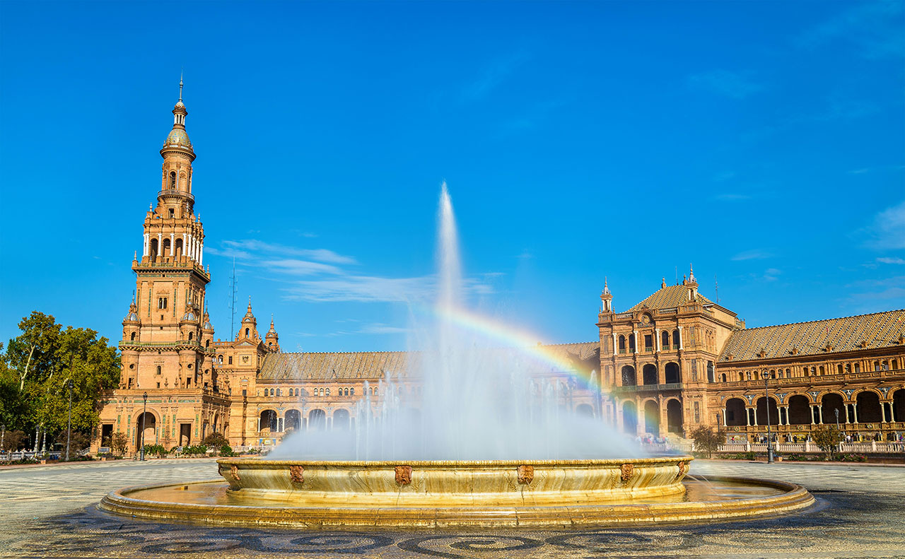 Plaza de España in Seville on a bright day with its large fountain creating a rainbow against the blue sky.