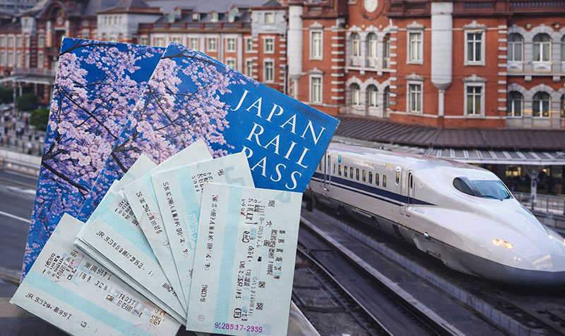 Hand holding multiple Japan Rail Passes in front of a Shinkansen bullet train at Tokyo Station