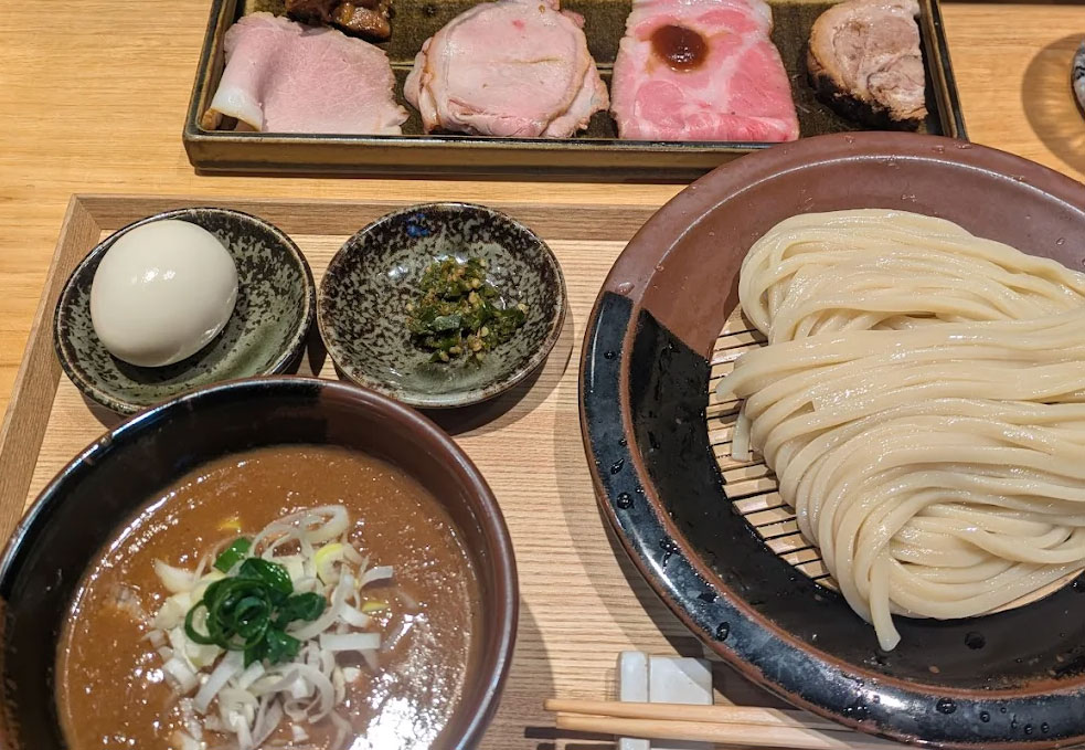 A bowl of thick, dark tonkotsu-gyokai dipping broth next to a plate of cold, thick wheat noodles topped with assorted chashu pork cuts and a soft-boiled egg from Ramen Tomita.