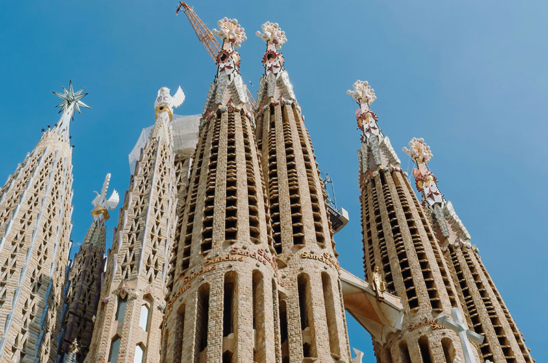 The intricate facade and towering spires of the Sagrada Familia in Barcelona, with construction cranes visible.