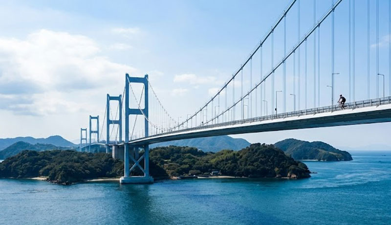 A cyclist riding across the massive Kurushima Kaikyo suspension bridge on the Shimanami Kaido route, with the deep blue Seto Inland Sea and green islands stretching out below.