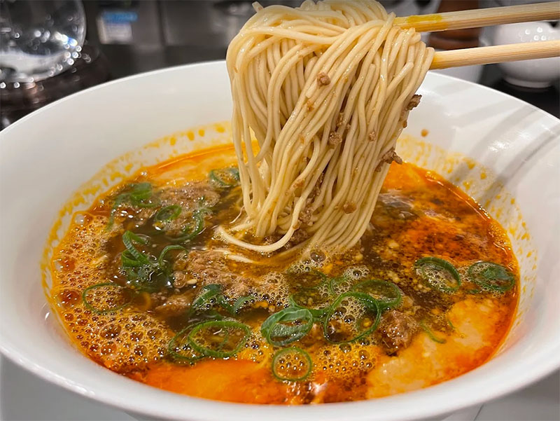 A bowl of Nakiryu's signature Tantanmen ramen featuring a rich, creamy orange sesame-based broth topped with minced pork, green onions, and a drizzle of vibrant red chili oil.