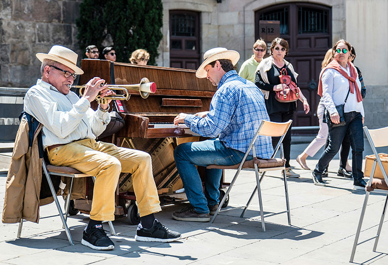 Street musicians playing trumpet and piano in a cathedral square, with pedestrians walking past.