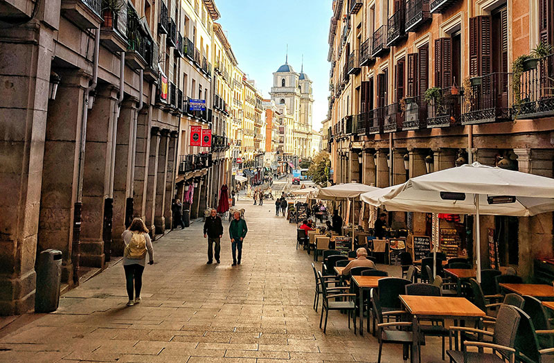 A street near Plaza Mayor in Madrid, lined with buildings and outdoor cafe seating, with pedestrians.