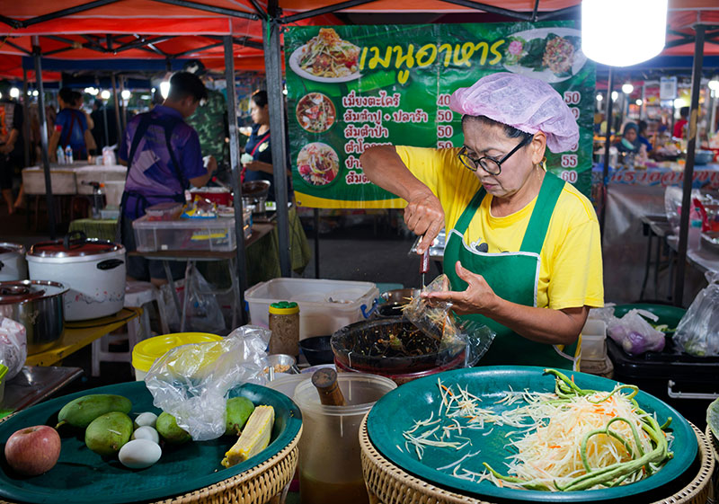 Thai street food vendor mixing fresh Yum Mamuang (spicy green mango salad) in a metal bowl at a street stall