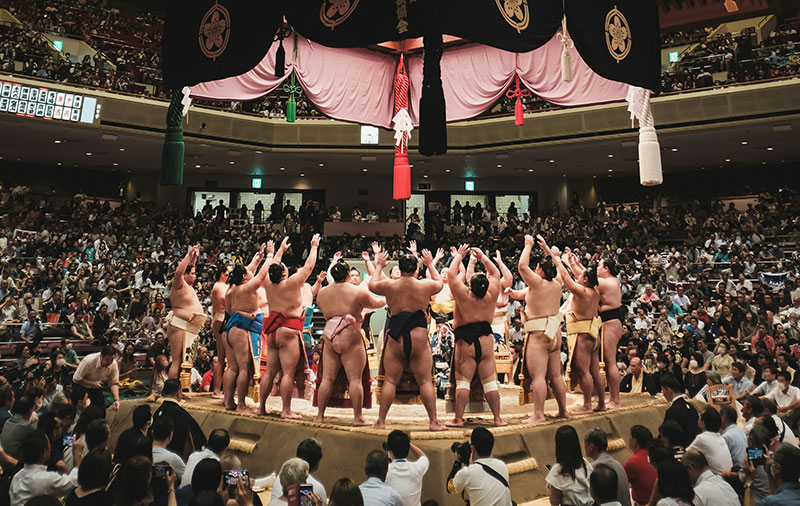 A wide-angle view of a professional sumo tournament at the Ryogoku Kokugikan, showing dozens of rikishi standing in the dohyo during the opening ceremony.