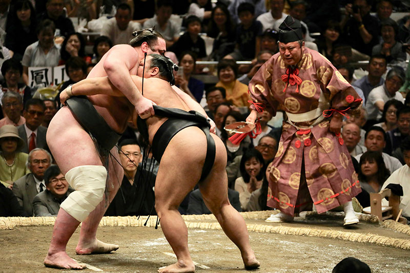 Two sumo wrestlers (rikishi) locked in a powerful grapple during a match in a traditional clay ring (dohyo).