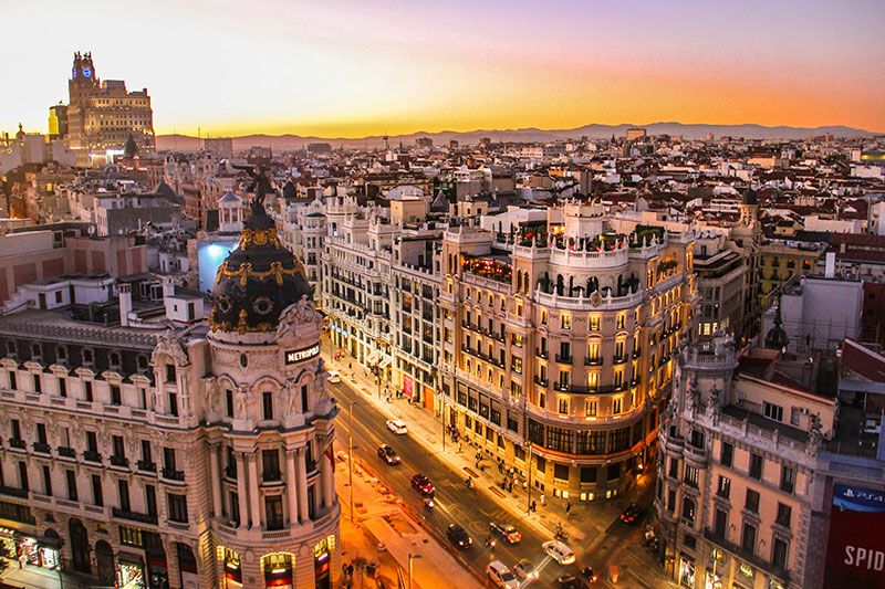 Elevated sunset view over Gran Vía in Madrid, with illuminated buildings and traffic on the street below.