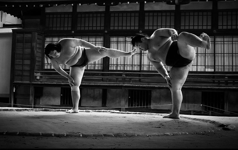 A black and white photo of two sumo wrestlers (rikishi) training outdoors, with one wrestler lifting the other in a demonstration of strength.