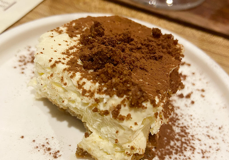 A slice of classic homemade Tiramisu served on a white plate in a Rome restaurant, showing layers of mascarpone cream and espresso-soaked ladyfingers dusted with cocoa powder.