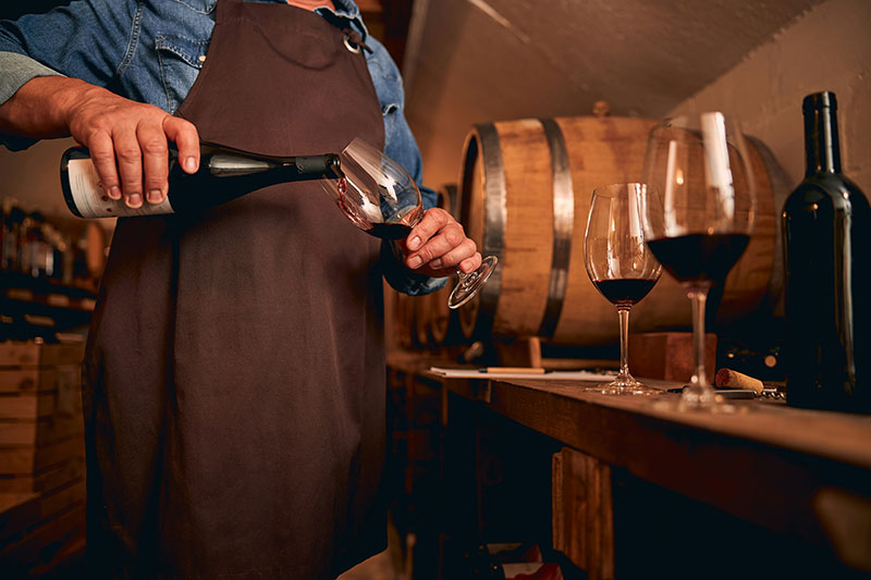 Sommelier pouring a glass of red Tuscan wine during a cellar tasting experience with wooden barrels in the background.