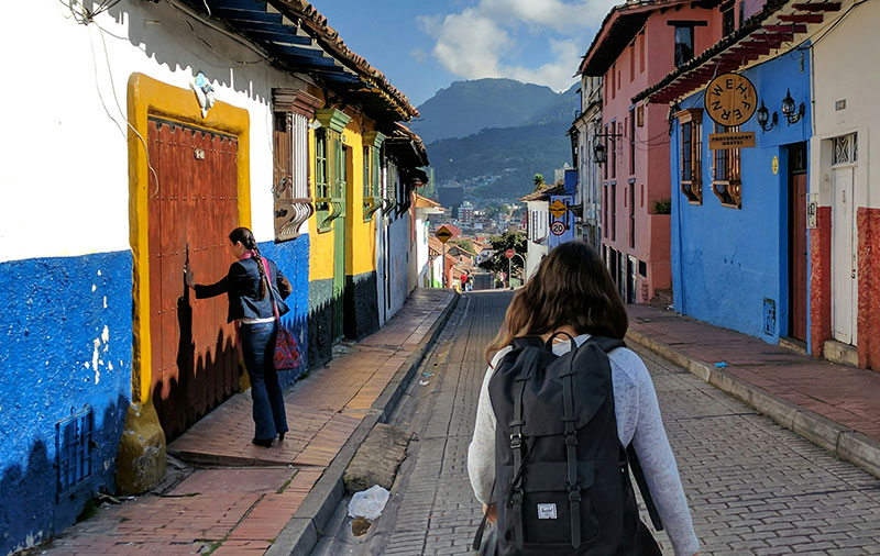 Woman walking down a colorful colonial street in La Candelaria, Bogota, Colombia, with mountains in the background.