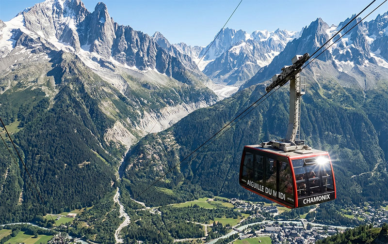 A photorealistic view of the red Aiguille du Midi cable car suspended high above the valley floor with the snowy Mont Blanc massif in the background.