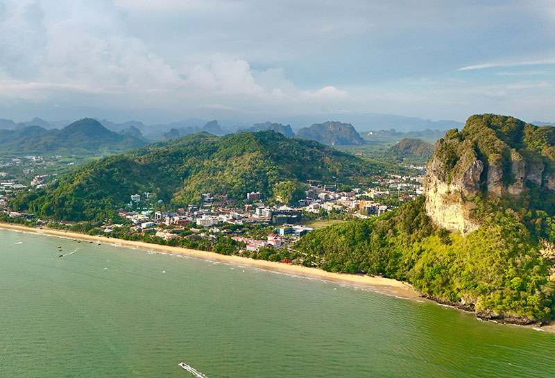 Aerial drone shot of Ao Nang Beach in Krabi, showing the long stretch of sand, the town nestled against green hills, and limestone cliffs.