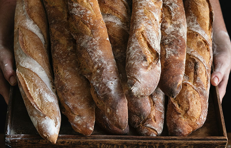 A baker holding a wooden tray stacked high with fresh, crusty French baguettes and rustic sourdough loaves.