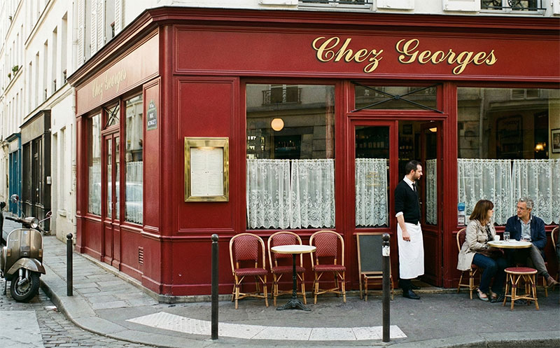 A classic, red-fronted Parisian bistro called Chez Georges on a quiet cobblestone street, with a waiter standing at the door and locals sitting on the small terrace, illustrating an authentic hidden gem.