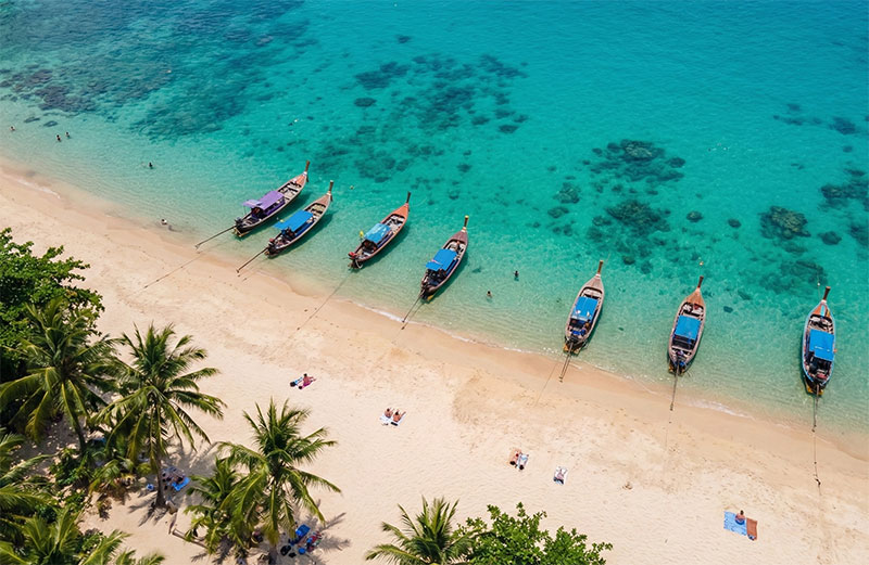 Aerial view of Banana Beach (Koh Hey) near Phuket featuring crystal clear turquoise water, traditional longtail boats anchored on white sand, and lush tropical greenery.