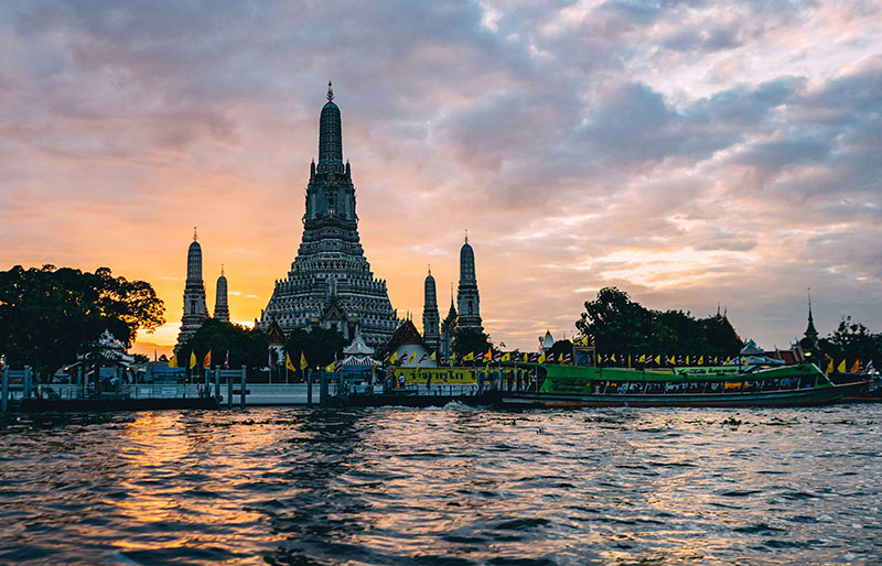Sunset view of the Wat Arun temple spire silhouetted against the sky on the Chao Phraya River in Bangkok.