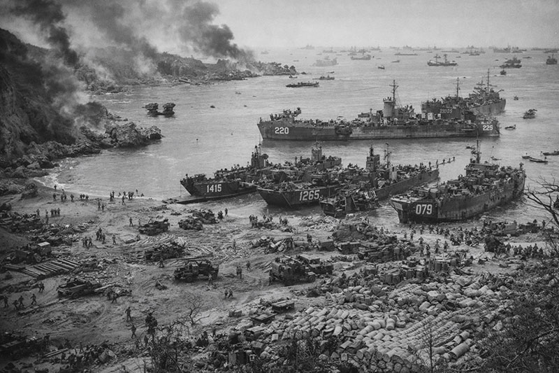 Aerial view of U.S. landing ships and troops unloading supplies on an Okinawa beach during the Battle of Okinawa in April 1945, with naval vessels offshore and smoke rising inland.