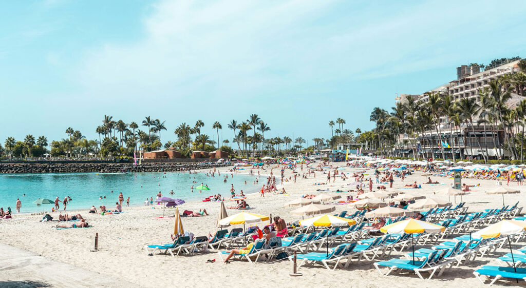 Crowded sunny beach with turquoise water and white sand under a clear blue sky.