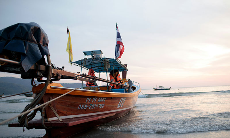 Close-up of a wooden longtail boat resting on the wet sand at sunset with a Thai flag waving from the stern.