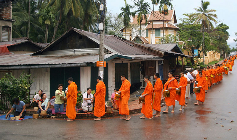 A procession of barefoot Buddhist monks wearing bright orange robes walking in a line down a wet street in Luang Prabang, Laos, while people kneel on mats along the sidewalk to place food offerings into their alms bowls during the morning Tak Bat ceremony.