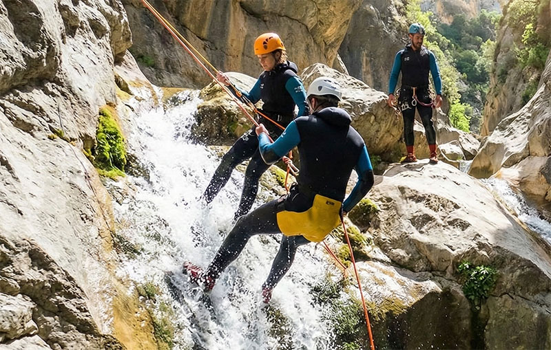 Adventurers canyoning in Sierra de Guara, Spain, with one person rappelling down a waterfall and another sliding down a natural rock chute into a turquoise pool.