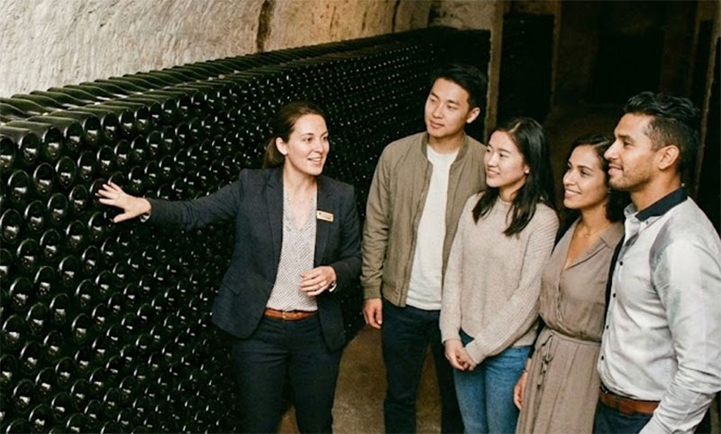 A tour guide in a chalk champagne cellar pointing to a wall of stacked bottles, explaining the aging process to a group of four young tourists.