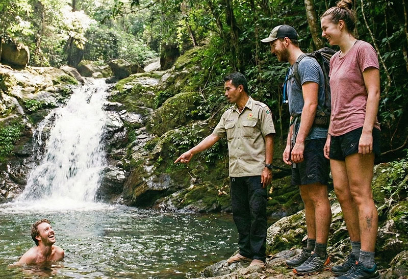 A group of friends on a day hike in the lush Chiang Mai jungle, laughing as one swims in a natural waterfall pool while a local Thai guide points out wildlife.