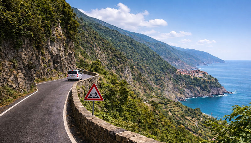 Narrow single-lane cliffside road overlooking a Cinque Terre village and the Ligurian Sea under a clear blue sky
