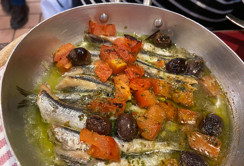 A silver pan filled with fresh anchovies cooked in olive oil with chopped tomatoes and black olives, a traditional dish of the Cinque Terre.