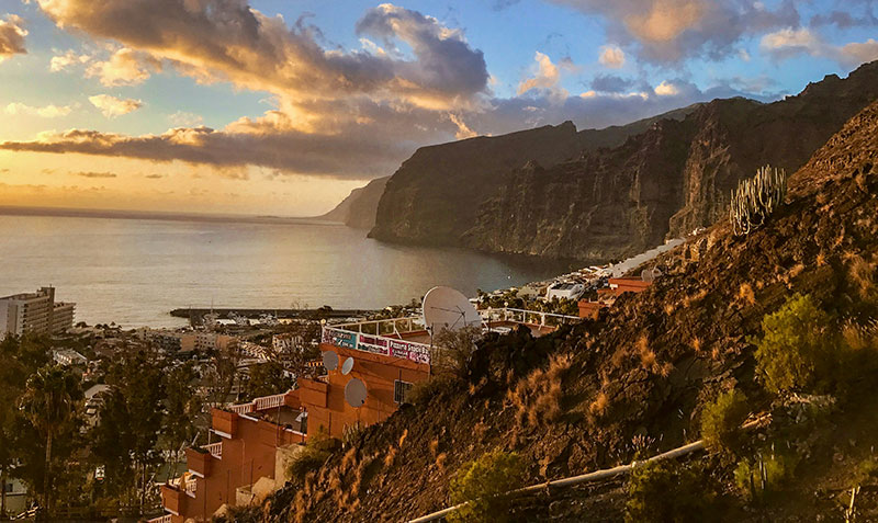 Aerial view of a coastal town in the Canary Islands with dramatic cliffs and a harbor at sunset.