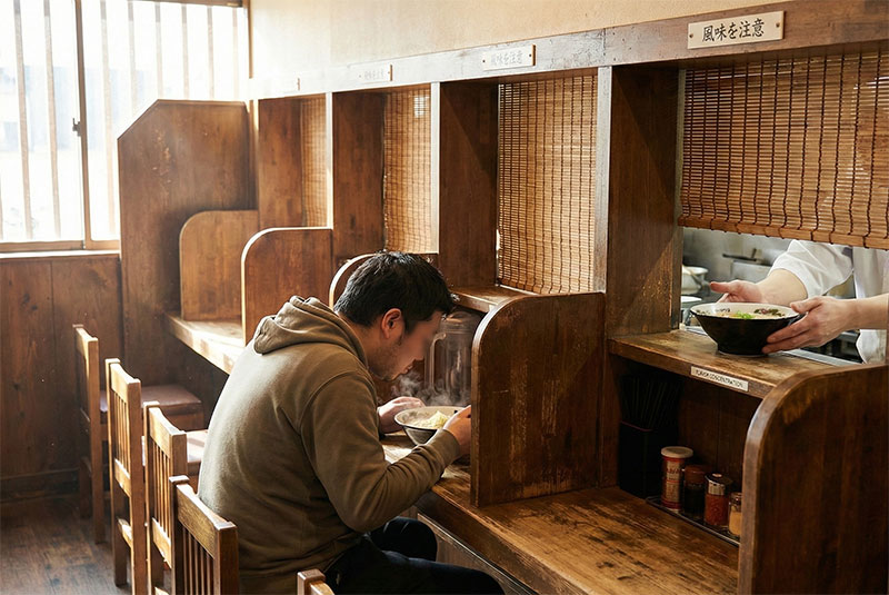 A solo diner enjoys a steaming bowl of ramen in a wooden partition booth filled with natural daylight, highlighting the rich broth and cozy atmosphere.